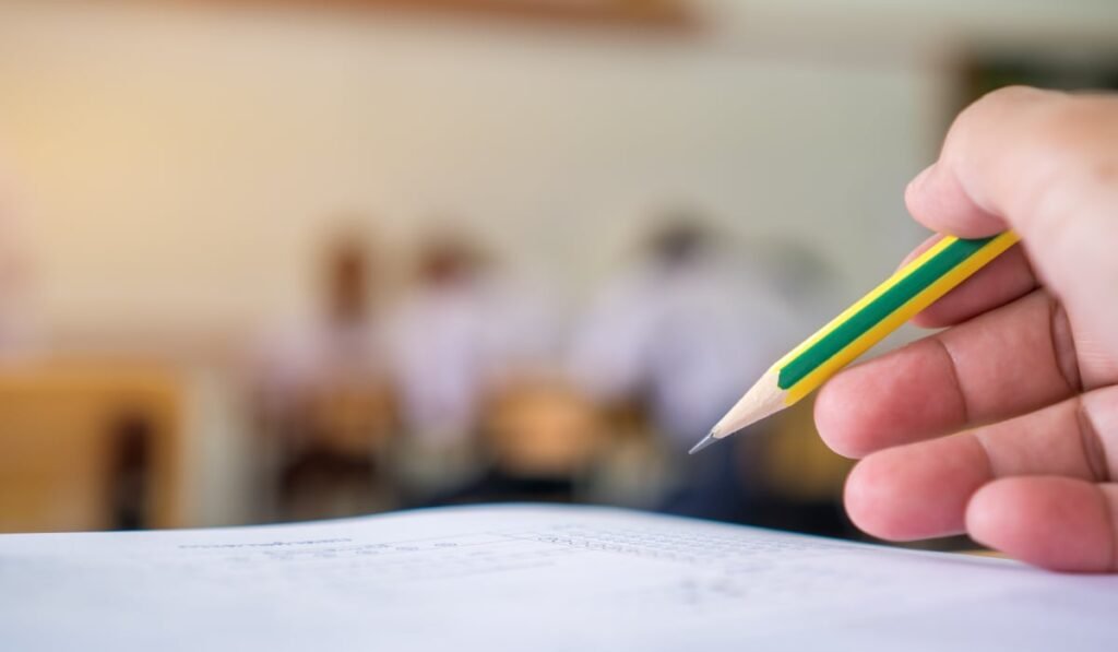 Imaginea arată o mână care ține un creion galben cu dungi verzi, pregătită să scrie pe o foaie, în timpul unui examen sau test, într-o sală de clasă. Fundalul este blurat, dar se disting alți elevi la bănci