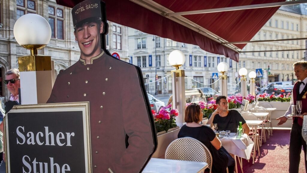 Imagine cu un bărbat și o femeie care stau la o masă a unei cafenele din Austria. În dreapta se află un ospătar care ține într-o mână o tavă cu pahare