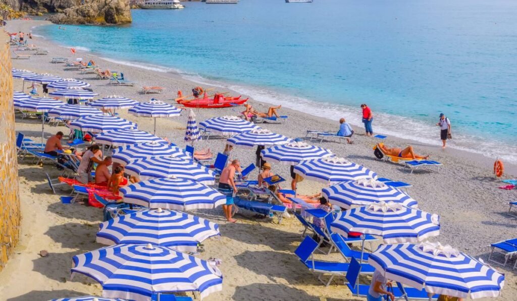 O plajă din Italia cu șezlonguri albastre și umbrele în dungi albastre cu alb, plaja nu este plină. Sunt doar câțiva oameni care fac baie în apa albastră a mării
