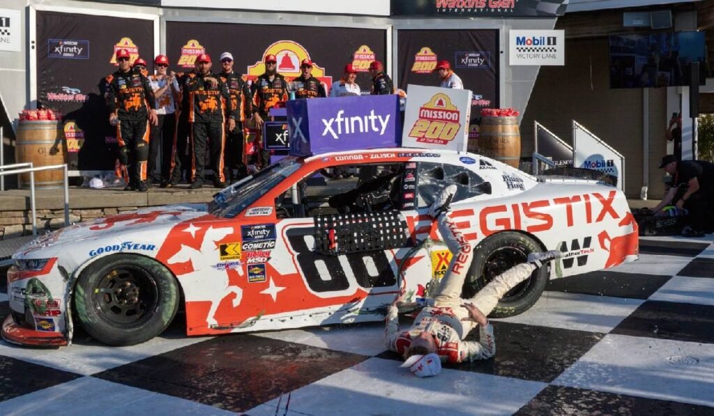 În imagine, Connor Zilish, prăbușit lângă Chevrolet-ul de pa care a căzut. El stă într-o poziție nefirească, întins pe asfalt, pe fundal sunt ceilalți concurenți NASCAR care-l privesc uimiți