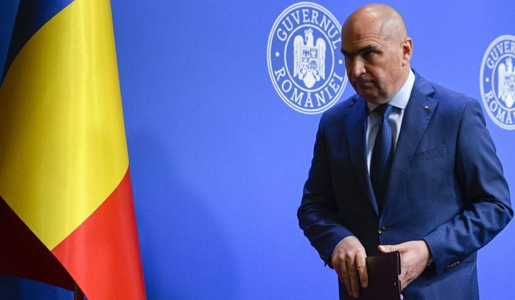 Premierul Ilie Bolojan, îmbrăcat în costum albadstru închis, pleaca la finalul unei conferinte de presa cu privire la reforma pensiilor in sitemul de Jutitie, marti, 29 iulie 2025, in Bucuresti. In fundal, în stânga se vede și steagul României.