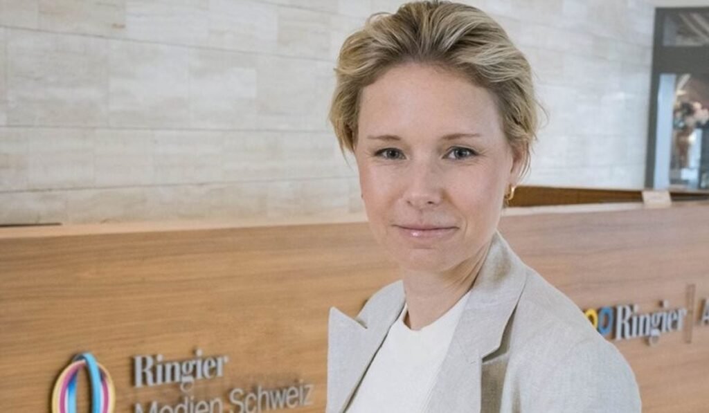 Ladina Heimgartner a fost aleasă vicepreședinte al Asociației Editorilor de Presă Elvețieni Ladina Heimgartner, îmbrăcată cu un sacou gri și o bluză albă, iar pe fundal un panou de lemn cu logo-ul Ringier