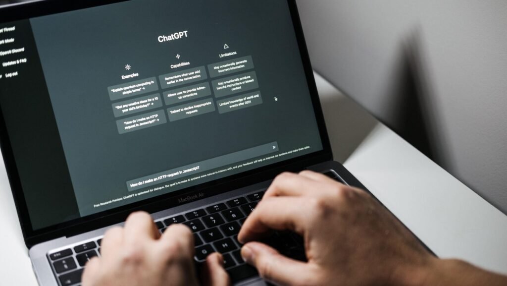 Imagine cu un laptop și cu mâinile unui bărbat pe tastatură. Pe ecranul dispozitivului este afișată aplicația ChatGPT