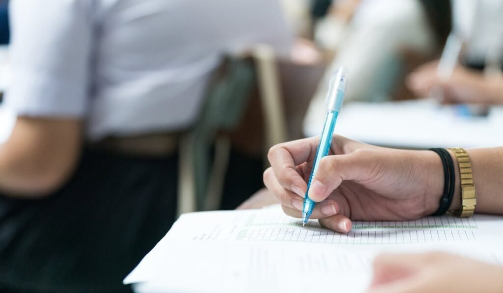 Imaginea arată o persoană care scrie cu un pix albastru pe o foaie, completând un test, într-un cadru ce pare a fi o sală de clasă sau de examen