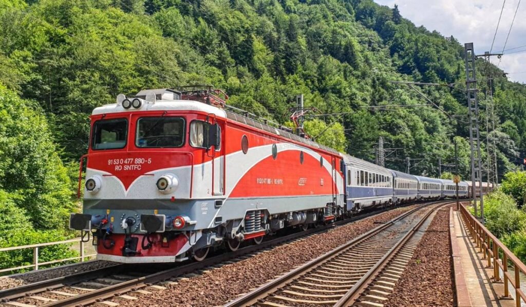 Un tren de călători cu locomotivă roșie merge pe calea ferată din România într-o zonă montană, este înconjurată linia de cale ferată de arbori cu frunze verzi.