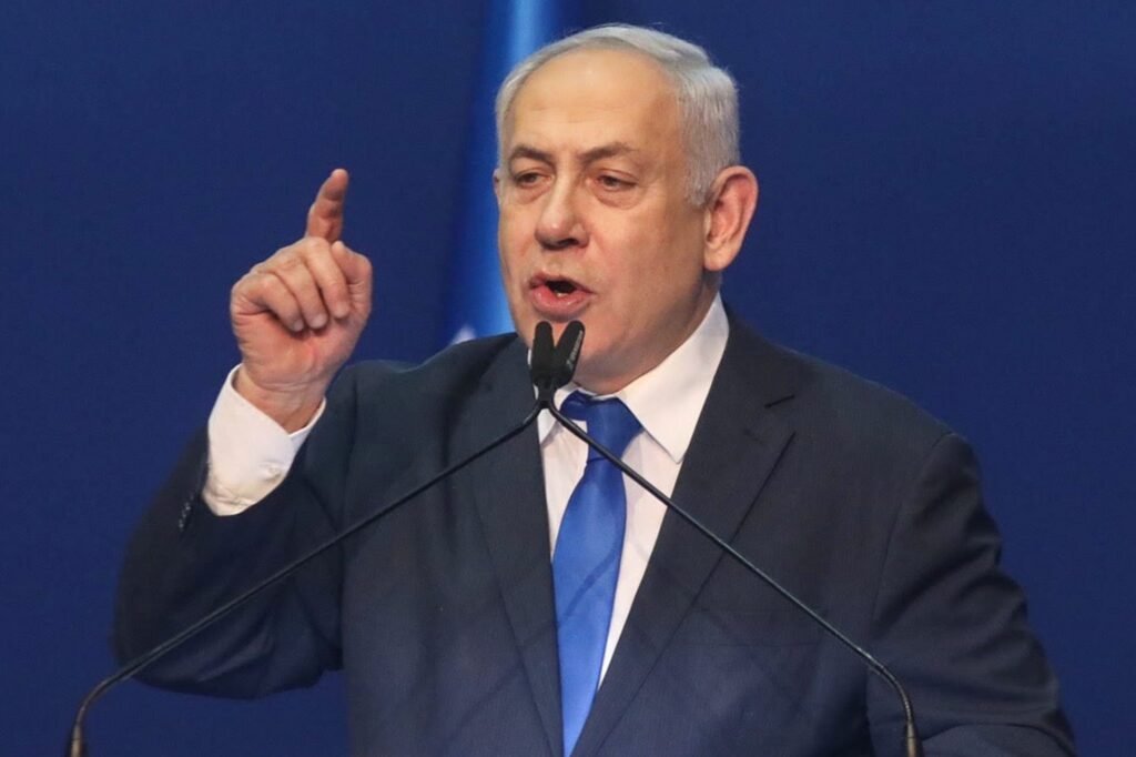 Benjamin Netanyahu, susține un discurs la microfon, purtând un costum bleumarin și o cravată albastră, ținând mâna dreaptă ridicată, cu degetul arătător ridicat
