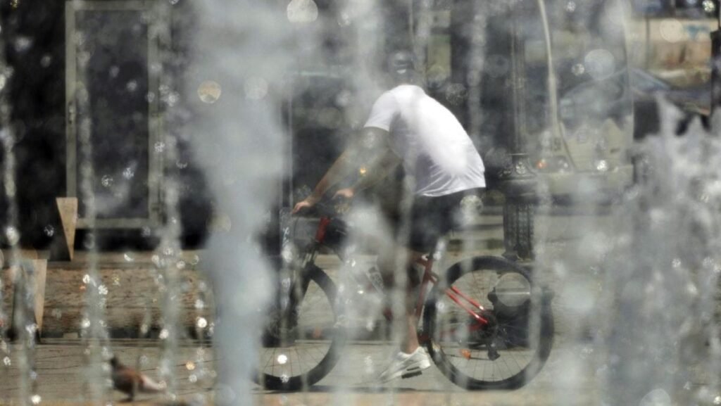 Un tânăr pe bicicletă, fotografiat cu spatele, în tricou alb și cu cască pe cap, trece prin dreptul unor fântâni din București, într-o zi caniculară de vară, București, 17 iulie 2025.
