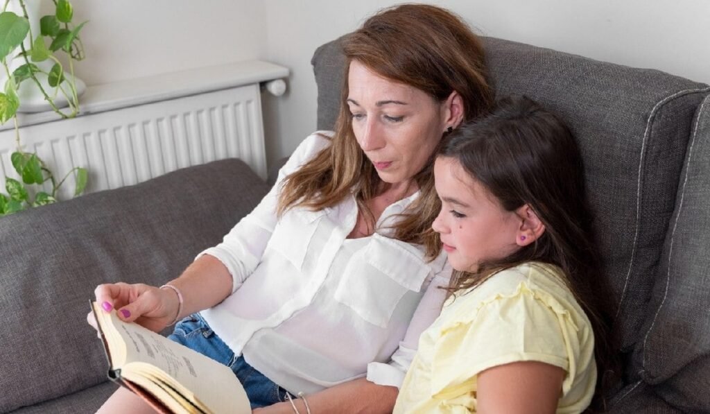 O mamă și o fiică fericite, așezate pe canapea și citind o carte.