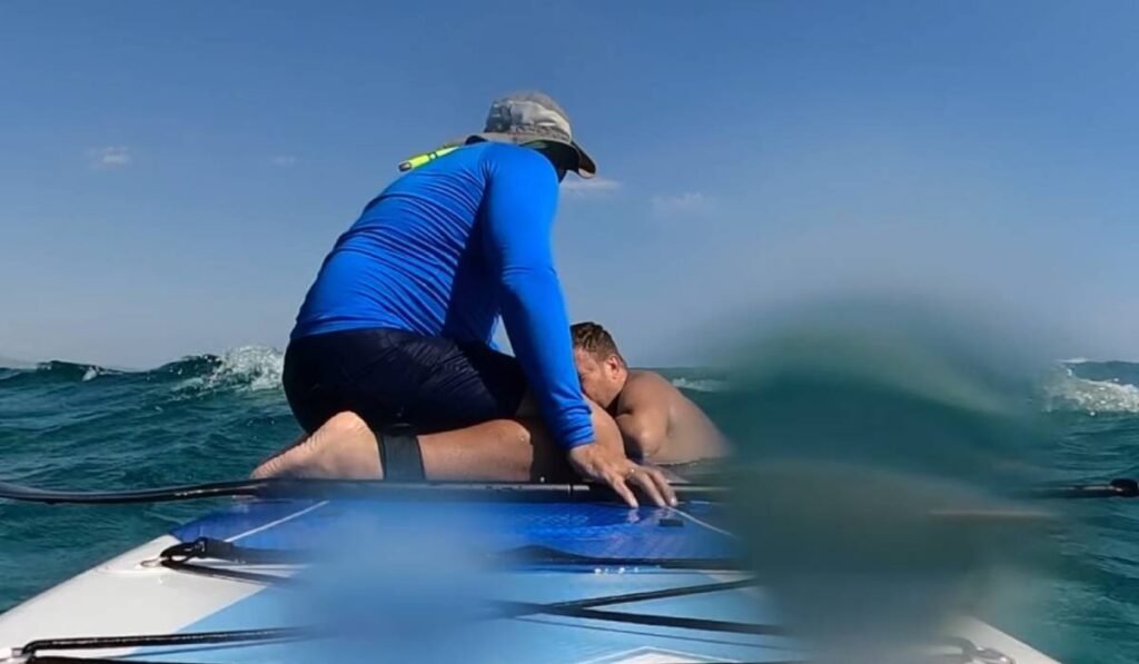 „Am auzit ţipetele copiilor” Momentul în care un român și cei doi copii au fost salvați de la înec în ultimul moment de un surfer care făcea paddle în Grecia Surferul grec care stă pe placa de SUP, în apă și se vede cum îi explică românului modul în care trebuie să se țină și să se agațe de placă