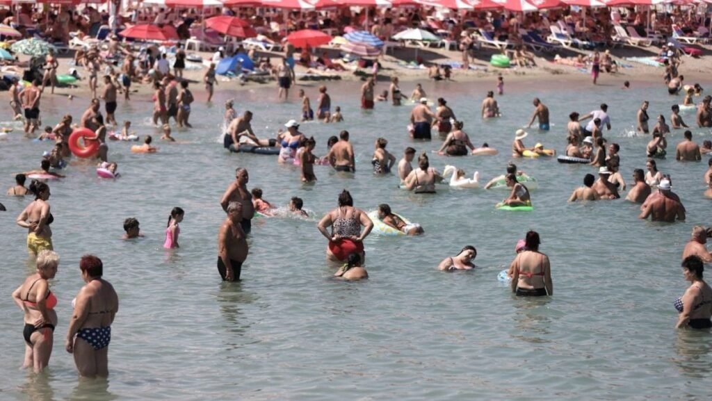 Mai mulți oameni în costume de baie sunt în apa mării, se văd și copii cu colace, iar în spate sunt umbrele mari de soare, roșii, unde turiștii stau la umbră