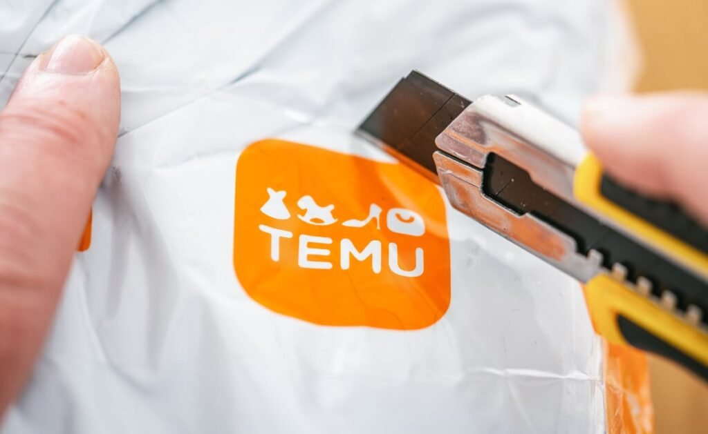 Imaginea surprinde de aproape o mână care ține un cutter cu mâner galben și negru, folosit pentru a deschide un colet alb, de la Temu. Logo-ul Temu, de culoare portocalie, este vizibil pe colet, alături de iconițe ce reprezintă articole vestimentare. Pachetul este pe cale să fie tăiat.