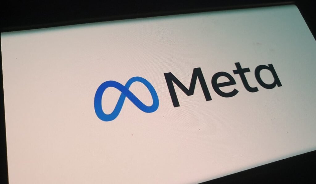 Imaginea arată sigla companiei Meta, afișată pe un ecran. Sigla este compusă din două elemente: Simbolul infinitului (o buclă orizontală) stilizat, de culoare albastră. Cuvântul "Meta" scris cu litere negre, cu un font modern, sans-serif, plasat lângă simbolul infinitului. Fundalul ecranului este de culoare albă, iar lumina emisă este ușor estompată, sugerând că ar putea fi o fotografie făcută într-un spațiu cu lumină redusă. Întregul ansamblu reprezintă logo-ul rebrenduirii companiei-mamă Facebook în Meta.