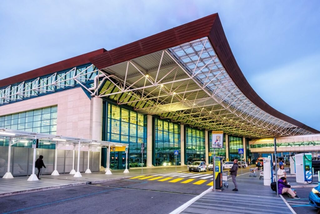 Imaginea surprinde intrarea modernă a unui aeroport la apus, cu o fațadă din sticlă și metal. Sub acoperișul curbat, câteva persoane așteaptă cu bagaje și cărucioare, în timp ce mașinile se deplasează pe banda de acces.