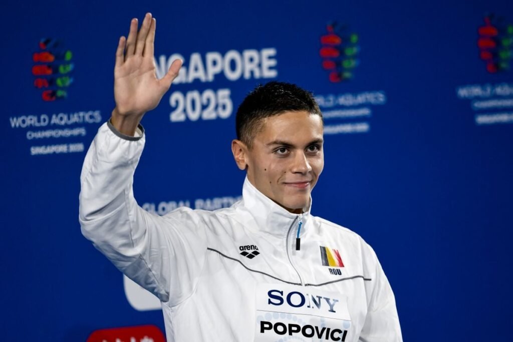 David Popovici salută publicul cu mâna dreaptă pe podiumul de premiere de la CM din Singapore, după ce a obținut medalia de aur în proba de 100 metri liber, iulie 2025