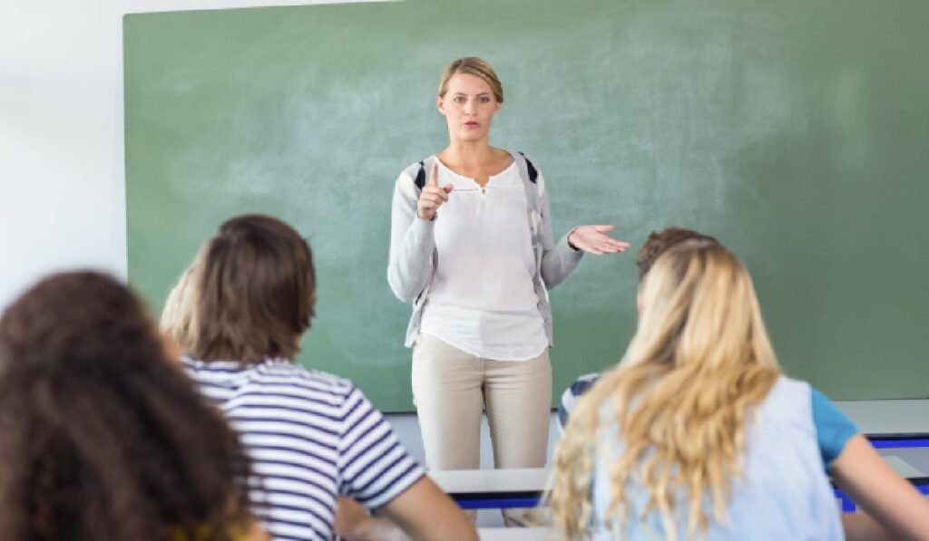 Imaginea prezintă o profesoară stând în fața unei clase de elevi. Ea este îmbrăcată casual, cu o cămașă albă, un cardigan gri deschis și pantaloni bej. Are părul blond, prins, și o expresie facială serioasă, cu o mână întinsă și un deget ridicat, ca și cum ar explica sau accentua un punct important. Cu cealaltă mână, face un gest deschis, sugerând o discuție. În fundal se află o tablă de culoare verde, iar în prim-plan, se văd din spate capetele elevilor, așezați la pupitre. Unii dintre ei au capetele întoarse, ascultând-o pe profesoară. Atmosfera generală a imaginii este de studiu și de predare-învățare, specifică unui mediu școlar.
