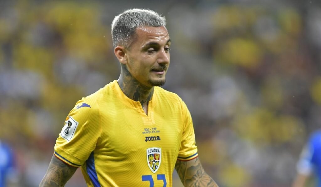 Imaginea este un prim-plan al fotbalistului Alexandru Mitriță. El poartă un tricou de joc al echipei naționale a României, de culoare galbenă, pe care se poate observa emblema Federației Române de Fotbal și un logo JOMA. Are o tunsoare scurtă, cu părul vopsit într-o nuanță deschisă de gri sau blond-cenușiu. O caracteristică vizibilă sunt numeroasele tatuaje care îi acoperă brațele și gâtul. Expresia sa facială este concentrată și serioasă.