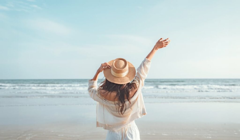 Imaginea arată o femeie pe o plajă, cu spatele la cameră. Ea poartă o pălărie de paie și haine ușoare, albe, care par a fi dintr-un material natural, cum ar fi inul. Femeia își ține brațele ridicate și pare să se bucure de moment, având o postură relaxată și fericită. În prim plan se vede nisipul umed al plajei și urmele valurilor care se retrag. Marea este calmă, cu valuri mici și blânde. La orizont, cerul albastru deschis se întâlnește cu apa, creând un peisaj senină. Lumina este moale, specifică unei dimineți sau după-amieze târzii.