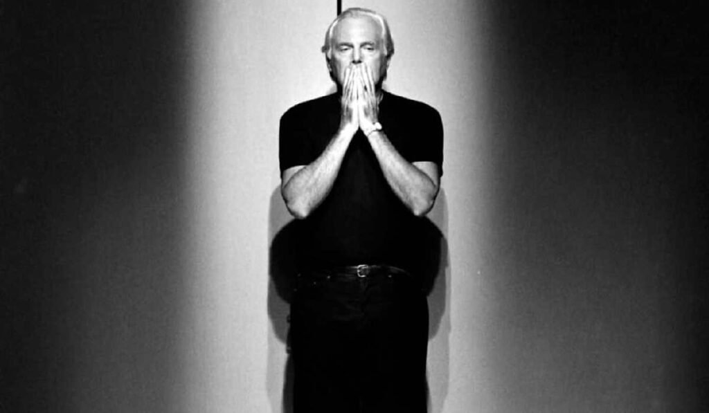 Creatorul de modă Giorgio Armani îmbrăcat în negru, ținând mâinile în jurul gurii