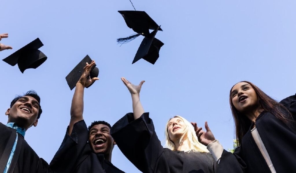 Absolvenții de facultate sărbătoresc aruncând în aer pălăriile și zâmbind fericiți în aer liber.