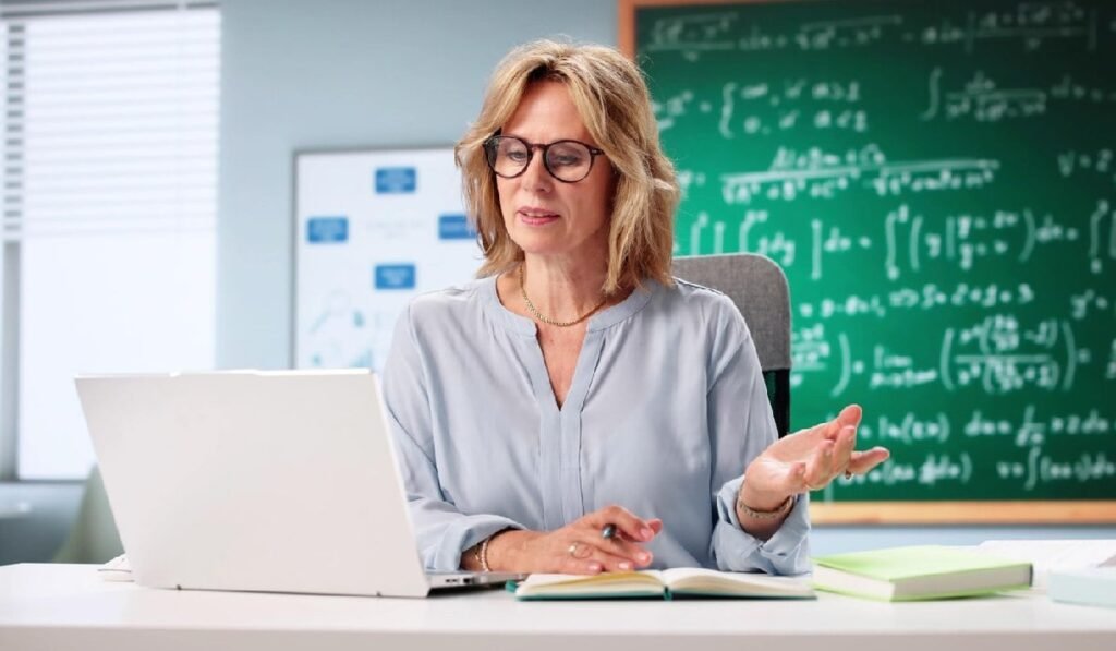 Un profesor de matematica foloseste laptopul pentru a preda, in spate este o tgabla verde, pe care este scris cu creta alba