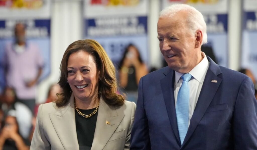 În imagine apar doi oficiali americani zâmbind și mergând unul lângă altul, îmbrăcați elegant, într-un cadru public. În fundal se văd bannere și câțiva participanți, sugerând că fotografia a fost surprinsă la un eveniment politic.