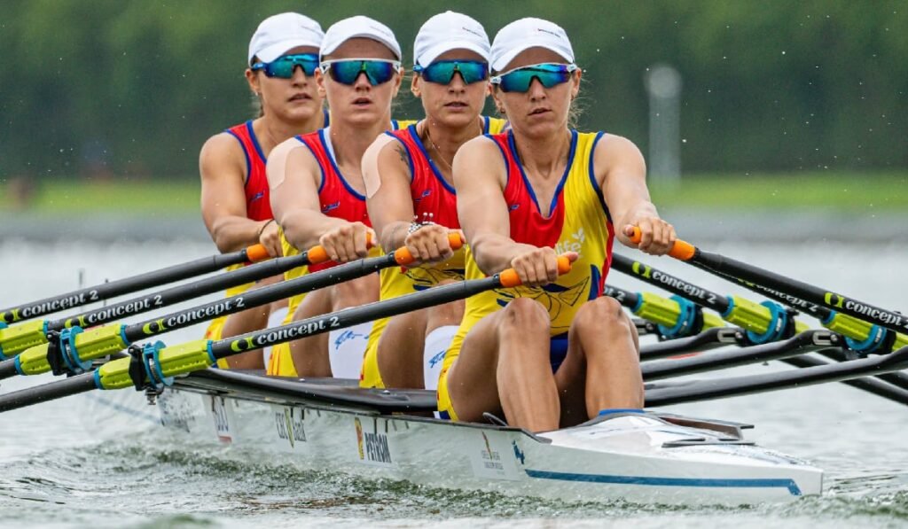 Imaginea surprinde un echipaj feminin de canotaj în timpul unei competiții. Cele patru sportive se află într-o barcă de tipul "patru vâsle", purtând uniforme de concurs de culoare galbenă și roșie. Toate au șepci albe și ochelari de soare. Atletele sunt într-un sincron perfect, vâslind cu multă forță și determinare. Expresiile faciale sunt concentrate, iar mușchii sunt încordați, sugerând efortul fizic intens. Barca se deplasează pe o suprafață de apă liniștită, pe care se văd reflexiile ușoare ale apei. Echipajul poartă uniforme ale echipei naționale a României. În partea de sus a imaginii, se observă un fundal estompat, de culoare gri-verde, care ar putea fi un peisaj cu arbori pe malul apei. Fotografia este focalizată pe mișcarea și efortul sportivelor.