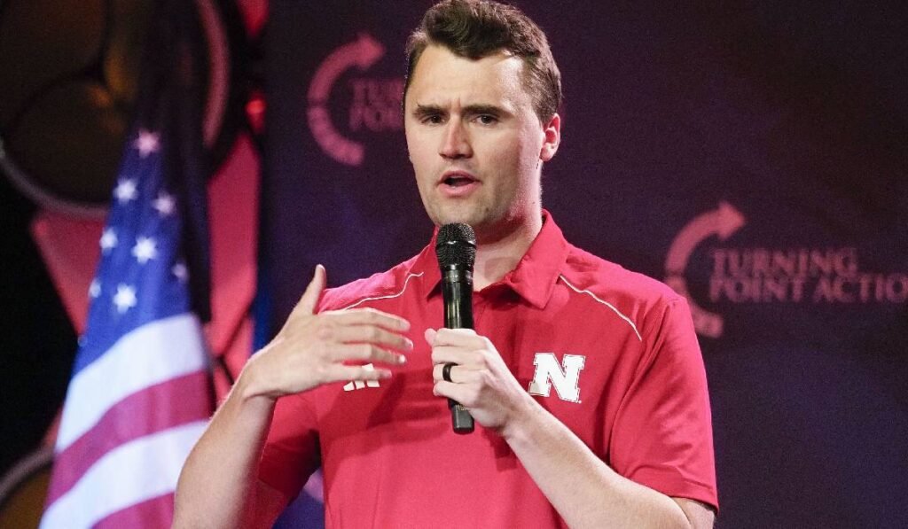 Charlie Kirk, îmbrăcat într-un tricou roșu, vorbește la microfon, pe fundal se vede un steag american