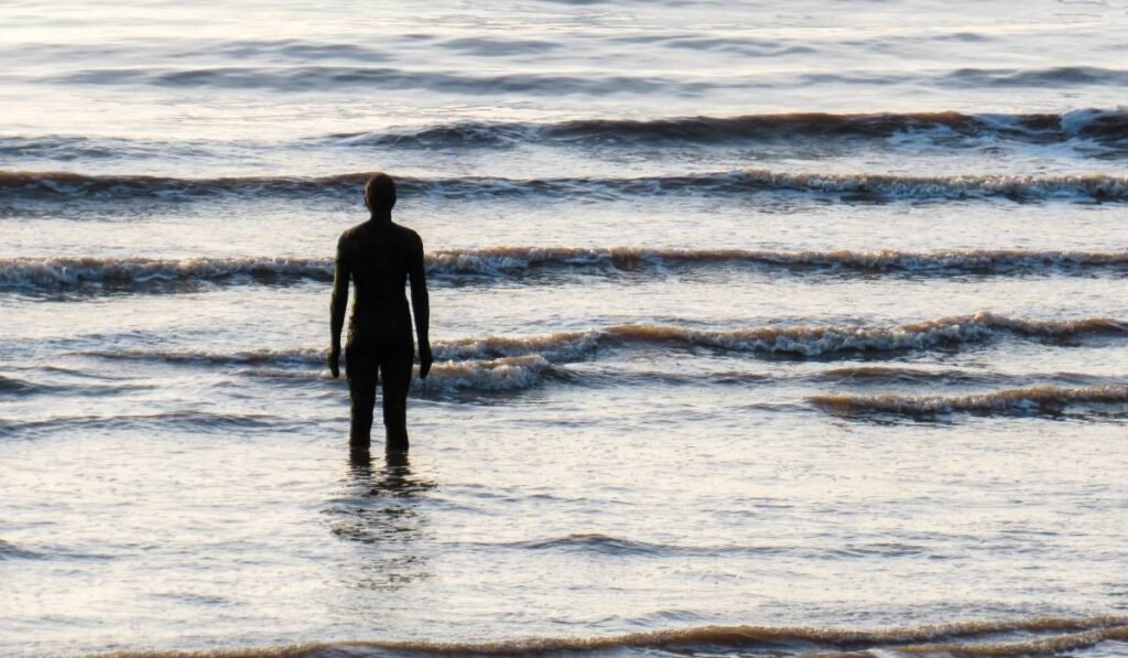 Un bărbat care stă la malul mării, apa îi vine până la genunchi și privește spre larg