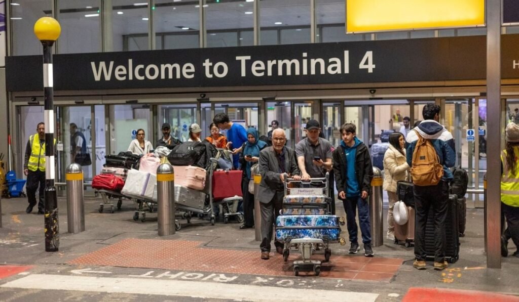 grup de oameni ieșind dintr-un terminal de aeroport, sub un panou mare cu inscripția „Welcome to Terminal 4”. Oamenii poartă haine diverse, de la casual la formal, și împing cărucioare pline cu bagaje de diferite dimensiuni și culori, inclusiv valize mari și pungi. În fundal, se văd ușile automate de sticlă ale terminalului și mai mulți oameni în interior, dar și doi angajați cu veste reflectorizante în partea stângă a imaginii