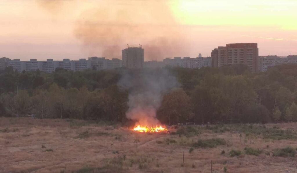 incendiu într-o zonă de vegetație uscată din zona retrocedată a parcului IOR din București. Un cerc de iarbă arde cu flăcări mari, vizibile, și degază fum gros și negru. pe fundal se vede peisajul urban al Bucureștiului, cu blocuri înalte iar pe fundal lumina soarelui denotă faptul că este apus