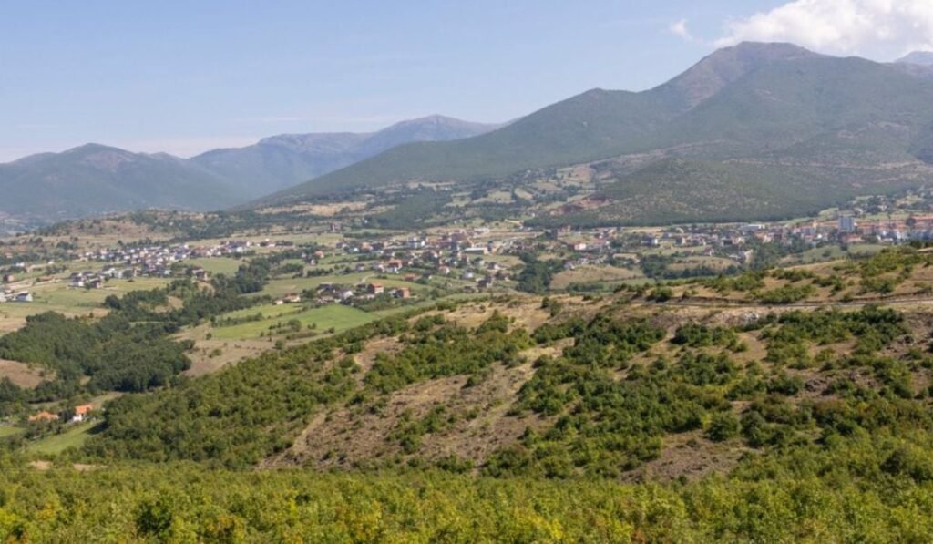 Satul albanez Golaj, vazut de la departare, pe dealuri cu iarba verde si paduri, in spate cu munti, este gol. 60% dintre locuitori au plecat în Marea Britanie, chiar și gangsterii