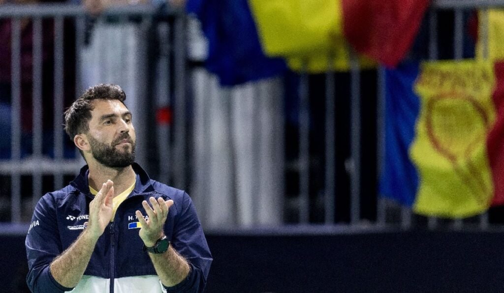 Horia Tecău, îmbrăcat cu o bluză de trening alb cu albastru, aplaudă în picioare, pe fundal fiind mai multe steeaguri tricolore românești, într-o tribună dintr-o sală de sport