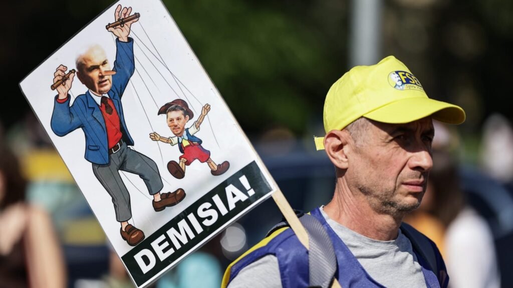 Imagine cu un protestatar care ține în mână o pancartă cu un afiș care-l înfățișează pe Ilie Bolojan drept păpușar, iar pe Daniel David drept o marionetă