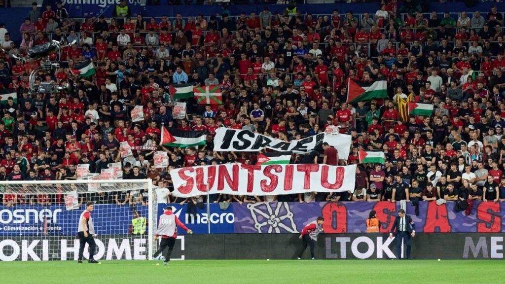 Ce mesaj a apărut pe bannerul anti-Israel și de ce au aruncat fanii mingi de tenis pe teren în LaLiga Imaginea surprinde un moment dintr-un meci de fotbal (posibil Osasuna-Getafe conform numelui fișierului), cu o tribună plină de suporteri îmbrăcați majoritar în roșu, care afișează diverse steaguri și bannere cu mesaje politice și locale. Printre steagurile palestiniene și basce, în centru se remarcă un banner mare cu textul "SUNTSITU" scris cu roșu pe fundal alb, sub un alt banner care pare să menționeze Israel.