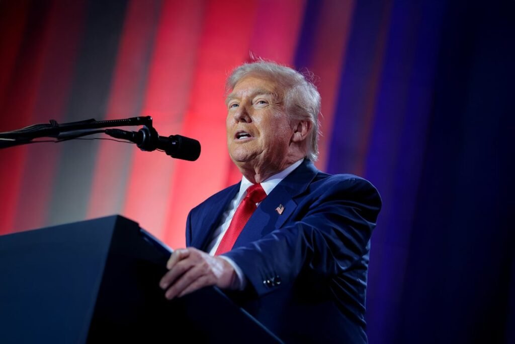 Donald Trmup imbrăcat intr-un costum bleumarin, completat de o cravată roșie, vorbește la un pupitru în fața unui microfon, fiind luminat puternic. În fundal se observă o scenă cu iluminare dramatică în culorile roșu, alb și albastru, sugerând că este un eveniment politic, un discurs sau o conferință.