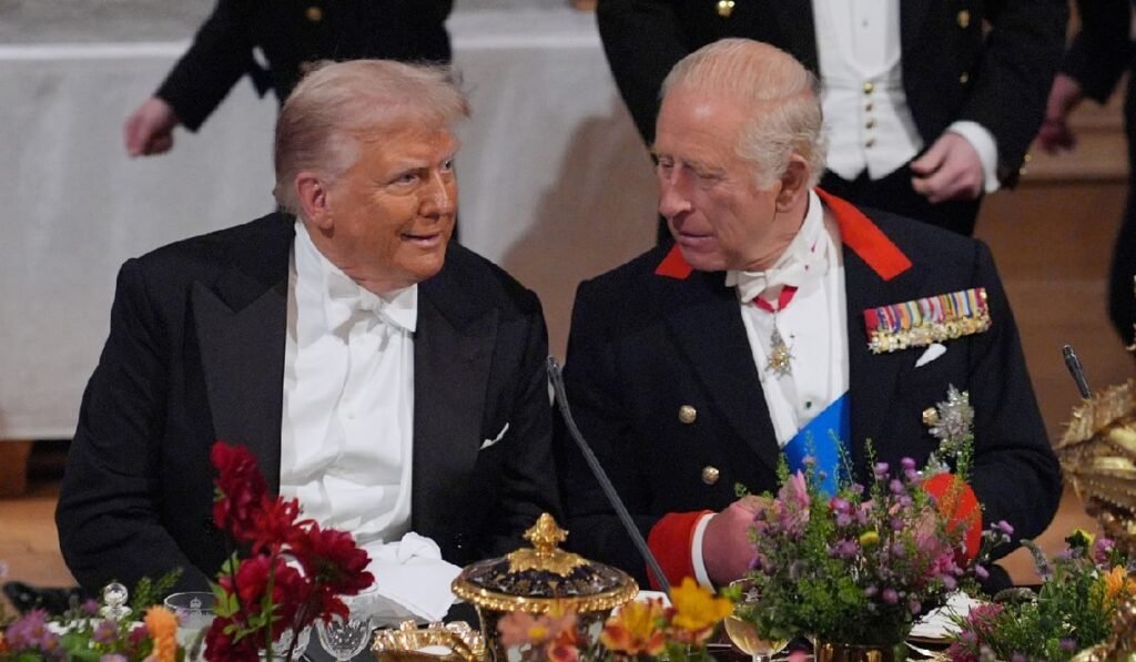 Imaginea surprinde o interacțiune formală între Donald Trump (stânga), îmbrăcat în frac cu papion alb, și Regele Charles al III-lea al Regatului Unit (dreapta), purtând o uniformă militară ceremonială cu medalii și eșarfă. Cei doi bărbați par să poarte o conversație animată la o masă festivă bogat decorată cu flori.