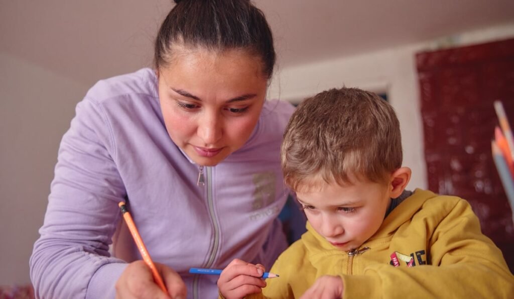 În câte comunități din România a ajuns proiectul „Start în educație” O mamă și fiul ei care sunt aplecați peste o masă și lucrează împreună la lecții. Amândoi folosesc creioane colorate, mama scriind pe hârtie în timp ce îl ghidează pe copil.