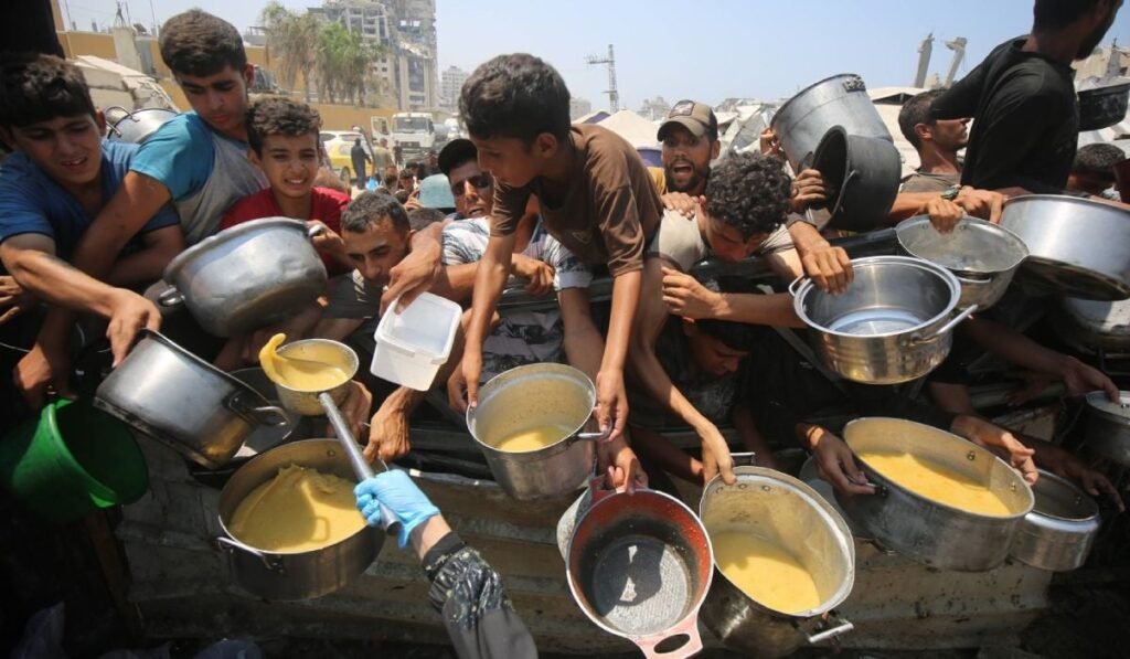 In imagine mai multi paliestinieni stau cu niste cratite intinse peste un grad ca sa primeasca mancare. Iulie, 2025