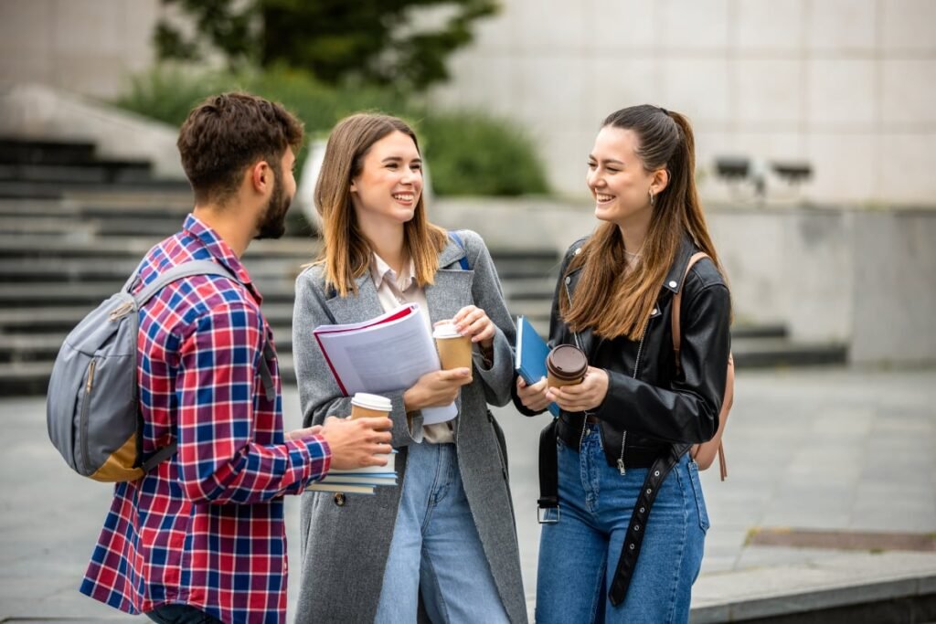 Imaginea surprinde trei studenți tineri care stau de vorbă și zâmbesc, ținând în mâini caiete și cești de cafea de carton, sugerând o pauză între cursuri. Ei sunt îmbrăcați casual, în haine moderne (blugi, sacouri și o jachetă de piele), cu fundalul exterior sugerând un campus universitar.