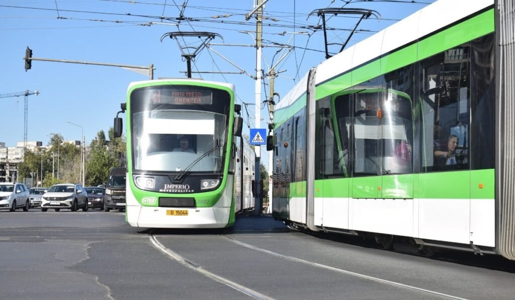 Imaginea, realizată într-o zi senină și luminoasă, surprinde două tramvaie moderne vopsite în alb și verde, care circulă pe șine paralele pe o stradă din oraș. În plan central, tramvaiul din stânga este marcat cu destinația "Piața Presei, Ghencea" (indicând probabil Bucureștiul) și are în fundal mașini la o intersecție.